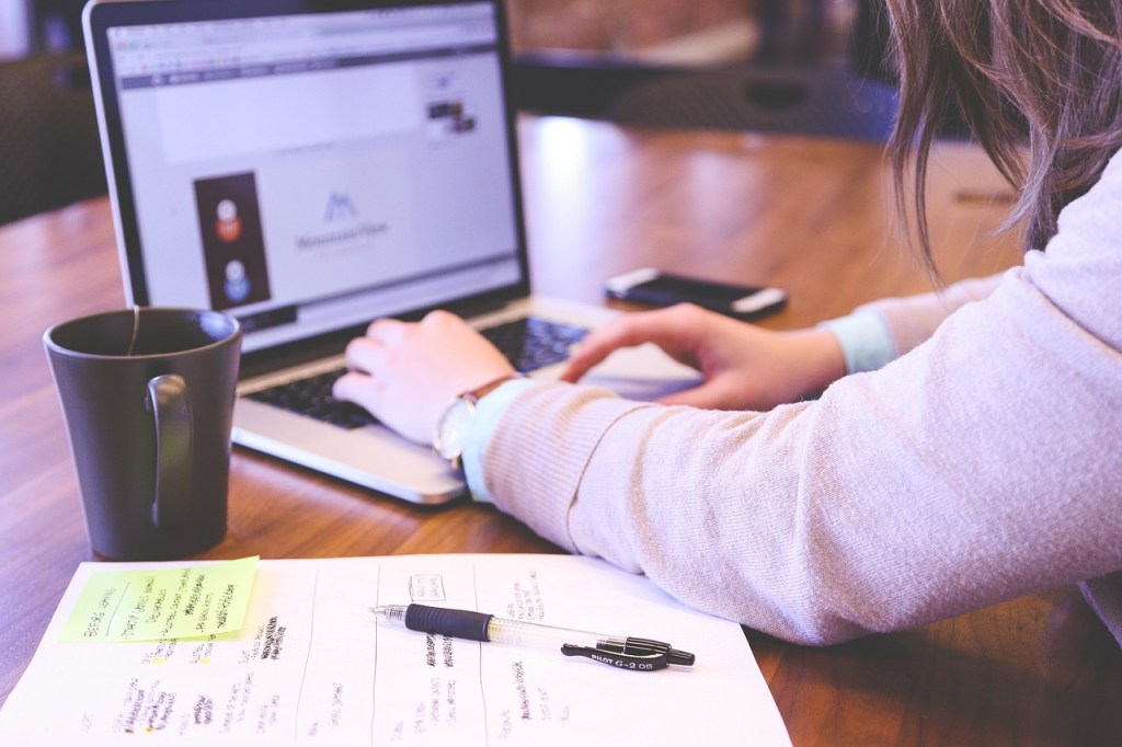 A woman sitting at a desk with a mug, a laptop, and a notebook with her. She is typing on her laptop, and only her arms are visible.